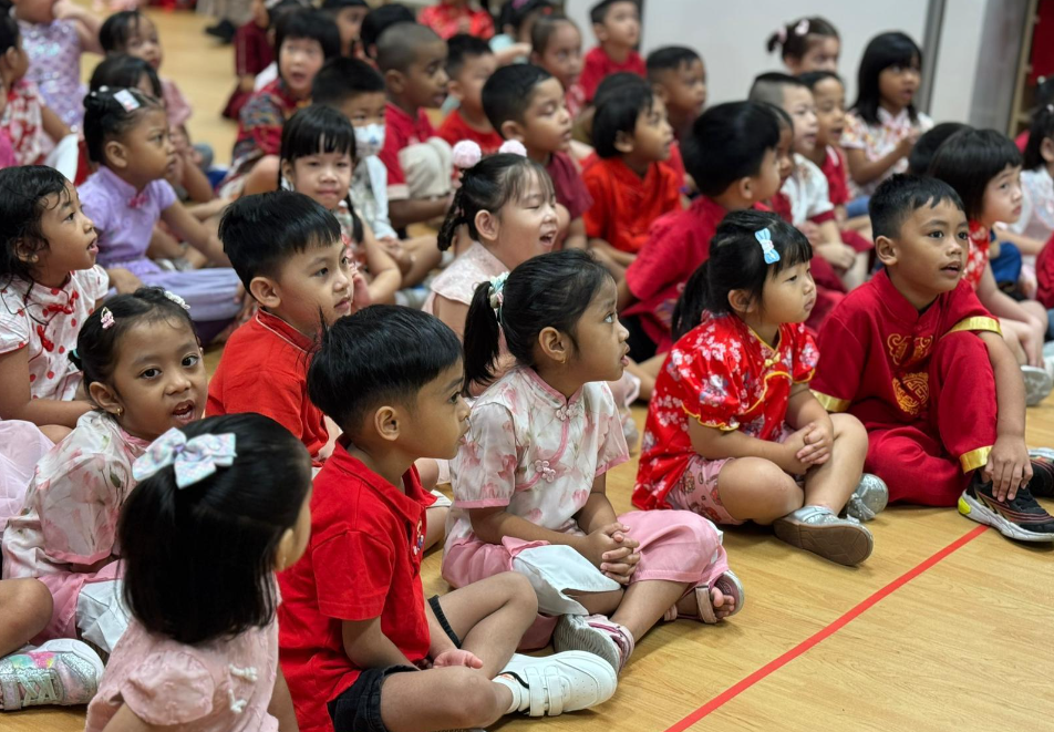 Group of young children seated on a gym floor, watching attentively, many in red and pink outfits.